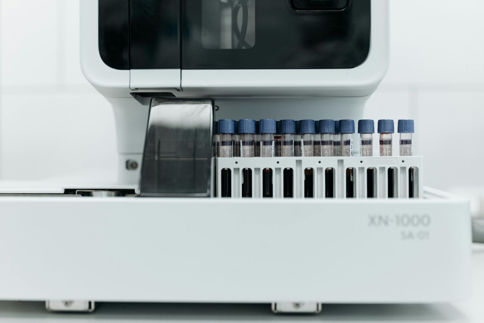 A laboratory analyzer machine holds multiple test tubes with blue caps arranged in a row on a white tray.